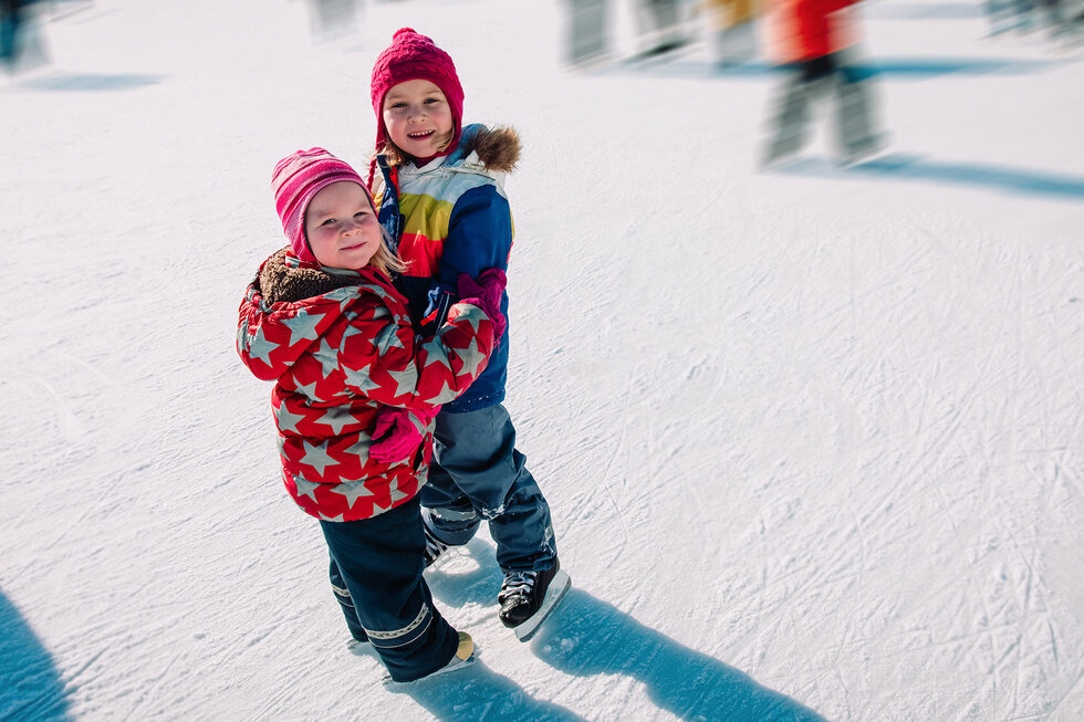 Zwei fröhliche Kinder stehen auf dem Eis, umarmt und lächelnd, während sie sich in bunten Winterjacken warmhalten.