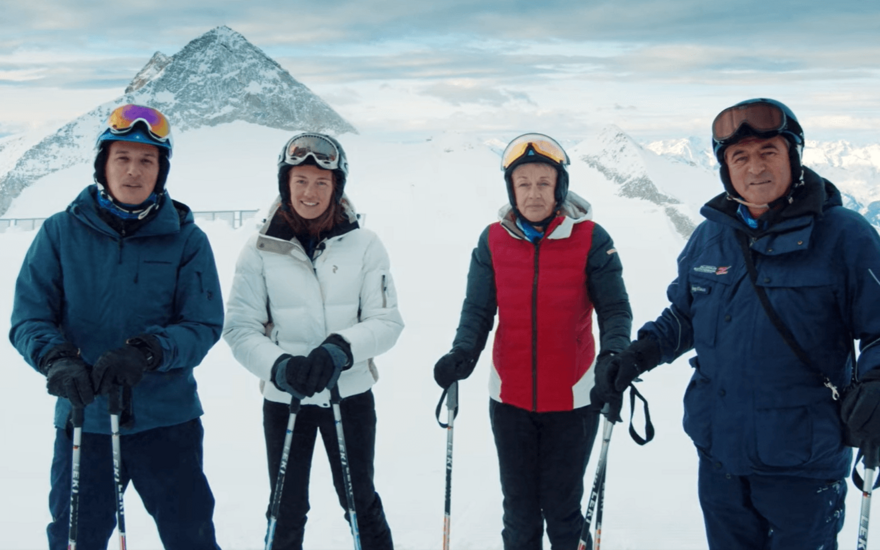 Vier Skifahrer stehen auf einer schneebedeckten Piste, umgeben von majestätischen Bergen und einem klaren Himmel.