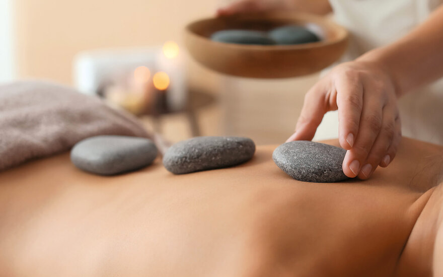 Eine Hand platziert einen glatten Stein auf dem Rücken einer Person, während eine andere Hand einen weiteren Stein hält.