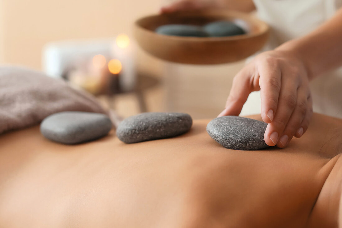 Eine Hand platziert einen glatten Stein auf dem Rücken einer Person, während eine andere Hand einen weiteren Stein hält.