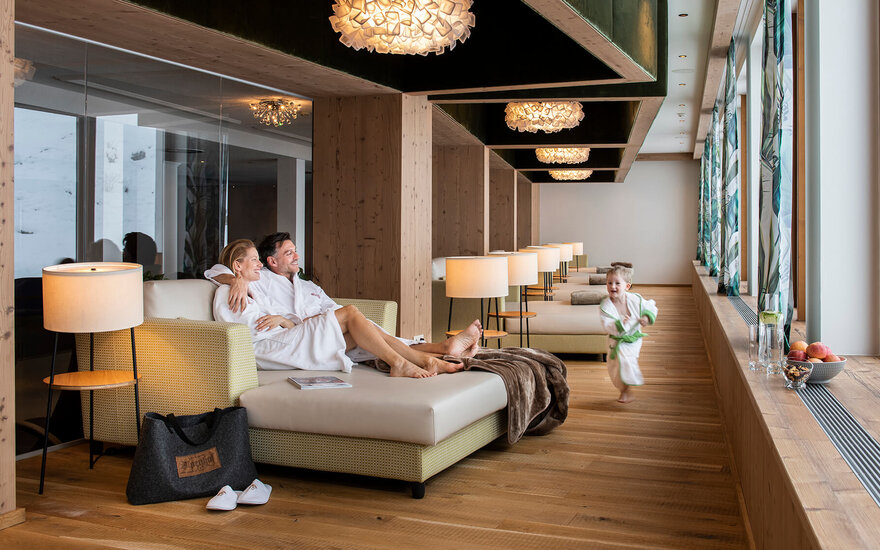 Ein Paar in Bademänteln entspannt auf einer Couch, während ein Kind fröhlich im Hintergrund läuft.