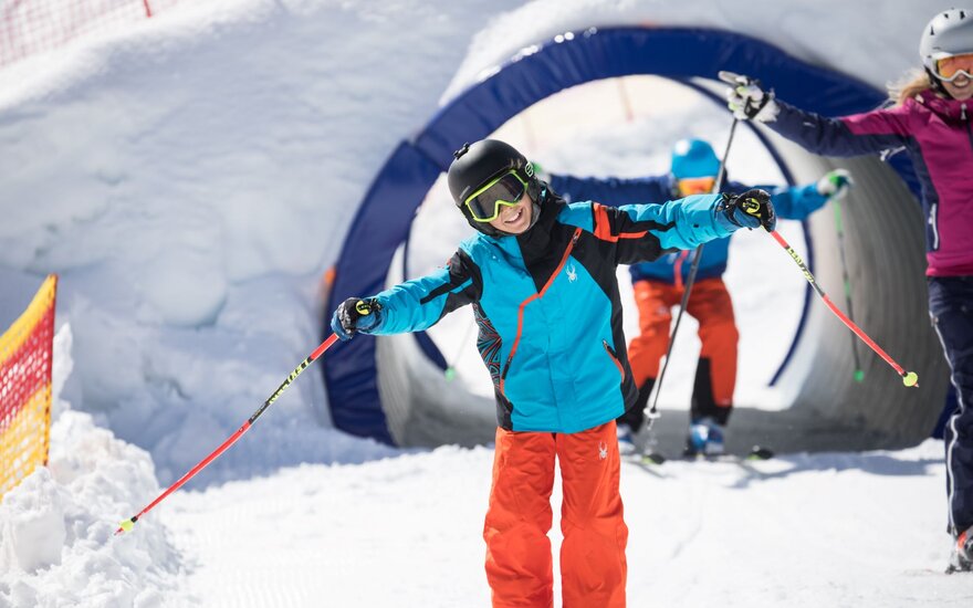 Ein fröhliches Kind in blauer und orangefarbener Skibekleidung genießt das Skifahren in einem verschneiten Skigebiet.