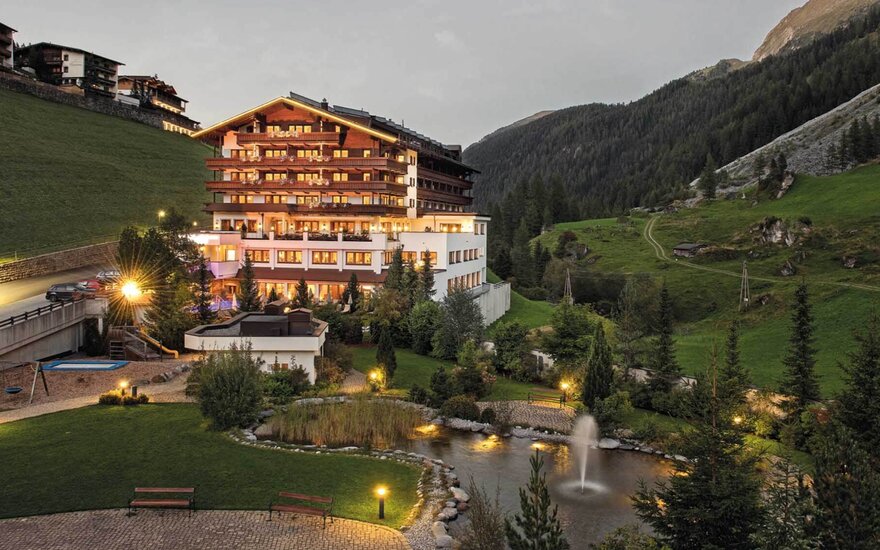 Ein elegantes Hotelgebäude mit warmem Licht, umgeben von grünen Hügeln und einem kleinen Teich mit einem Springbrunnen.