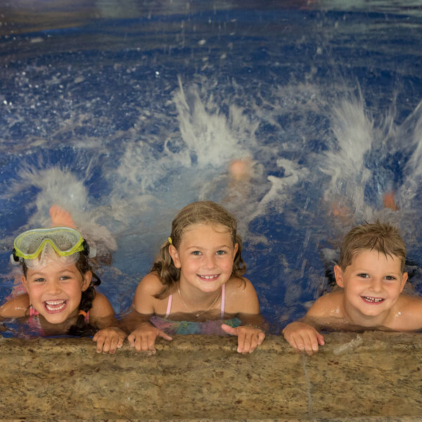 Drei fröhliche Kinder im Wasser, die am Beckenrand lächeln und mit Wasser spritzen, während sie Schwimmbrillen tragen.