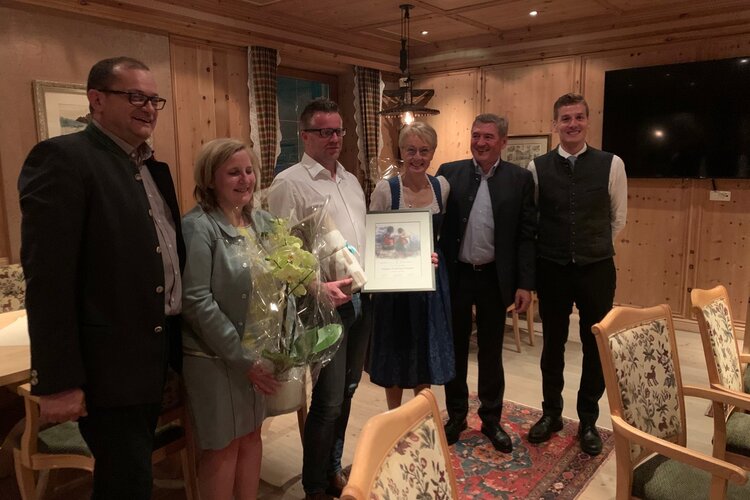 In einem warmen, holzverkleideten Raum feiern sechs Personen einen besonderen Anlass mit Geschenken und Lächeln.