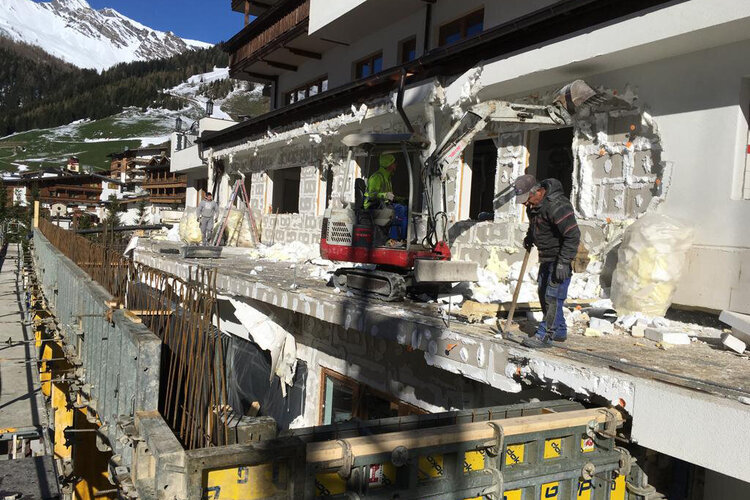 Auf einer Baustelle arbeiten Handwerker an der Außenseite eines Hauses, um die Isolierung zu entfernen, umgeben von malerischen Bergen.