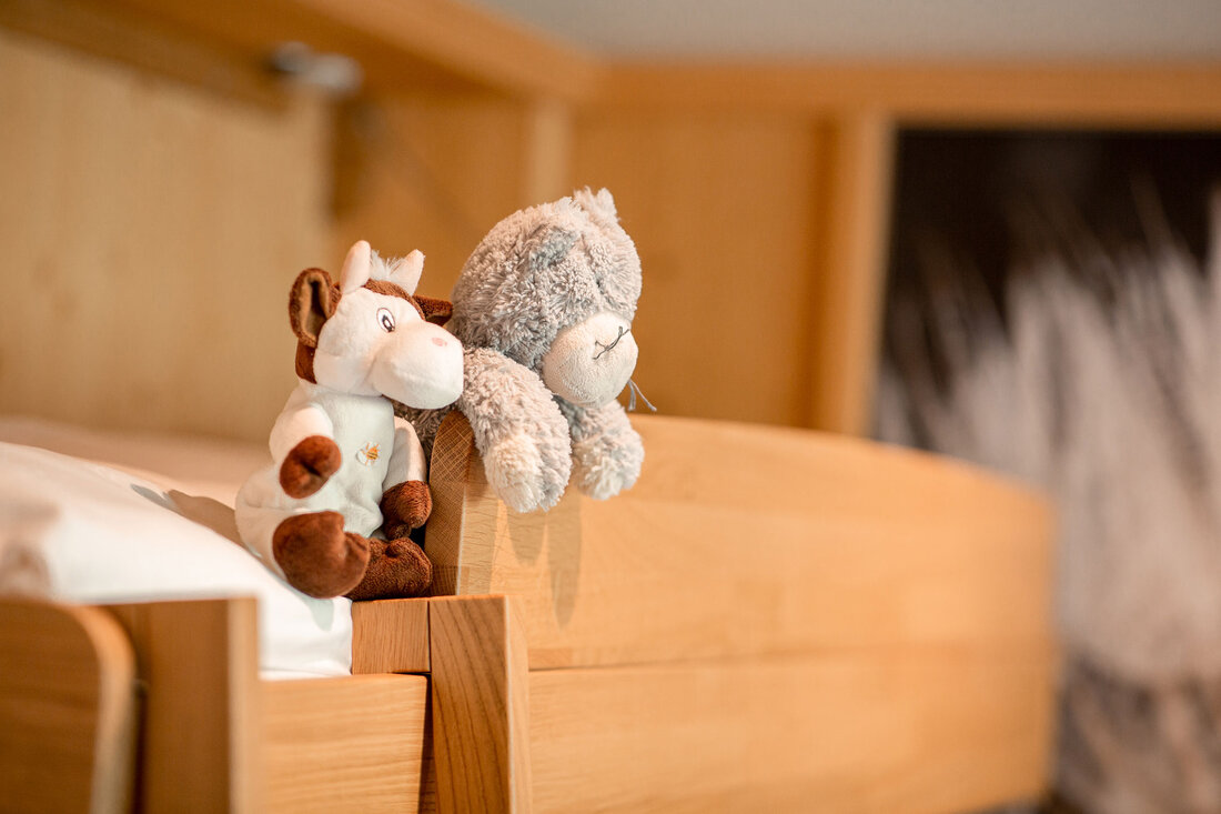 Zwei Kuscheltiere, ein Pferdchen und eine graue Katze, sitzen auf der Kante eines Holzbettes in einem gemütlichen Raum.