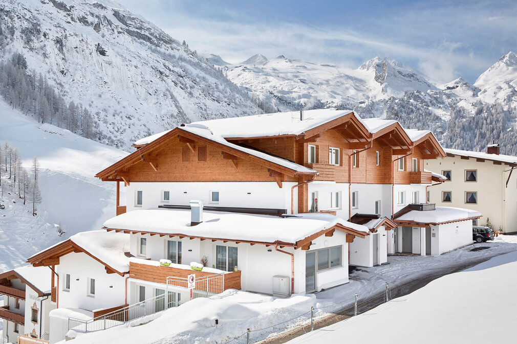 Eine moderne alpine Unterkunft mit Holzverkleidung, umgeben von schneebedeckten Bergen und klarem Himmel.