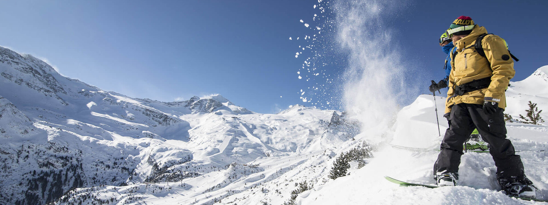 Zwei Snowboarder stehen auf einem schneebedeckten Hang, während Schnee in der Luft aufwirbelt und die Berge im Hintergrund sichtbar sind.