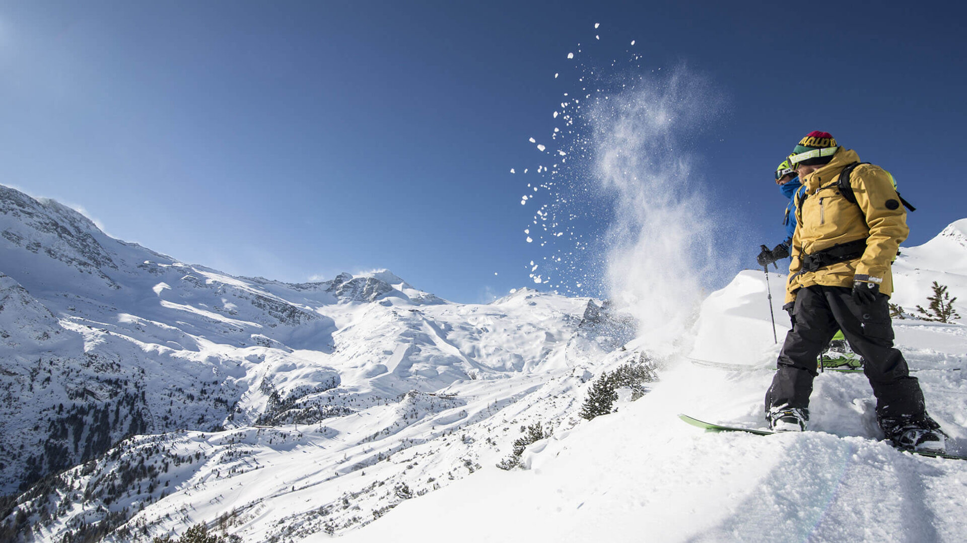 Zwei Snowboarder stehen auf einem schneebedeckten Hang, während Schnee in der Luft aufwirbelt und die Berge im Hintergrund sichtbar sind.