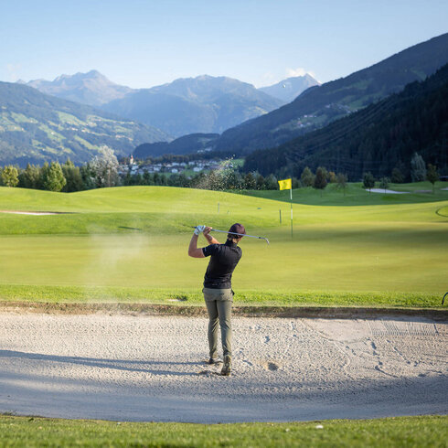 Ein Golfer schlägt aus einem Bunker heraus, während die Berge im Hintergrund eine malerische Kulisse bieten.