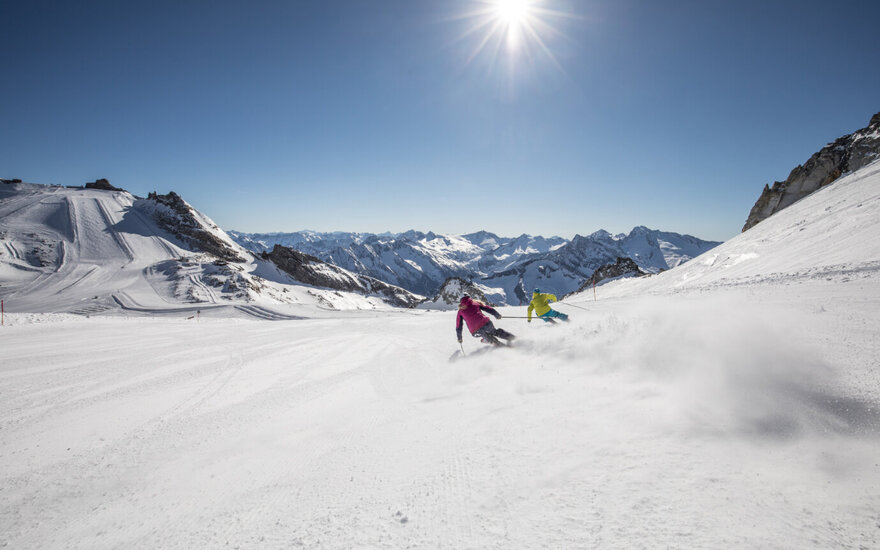 Zwei Skifahrer, einer in pink und der andere in gelb, gleiten über die schneebedeckte Piste mit majestätischen Bergen im Hintergrund.