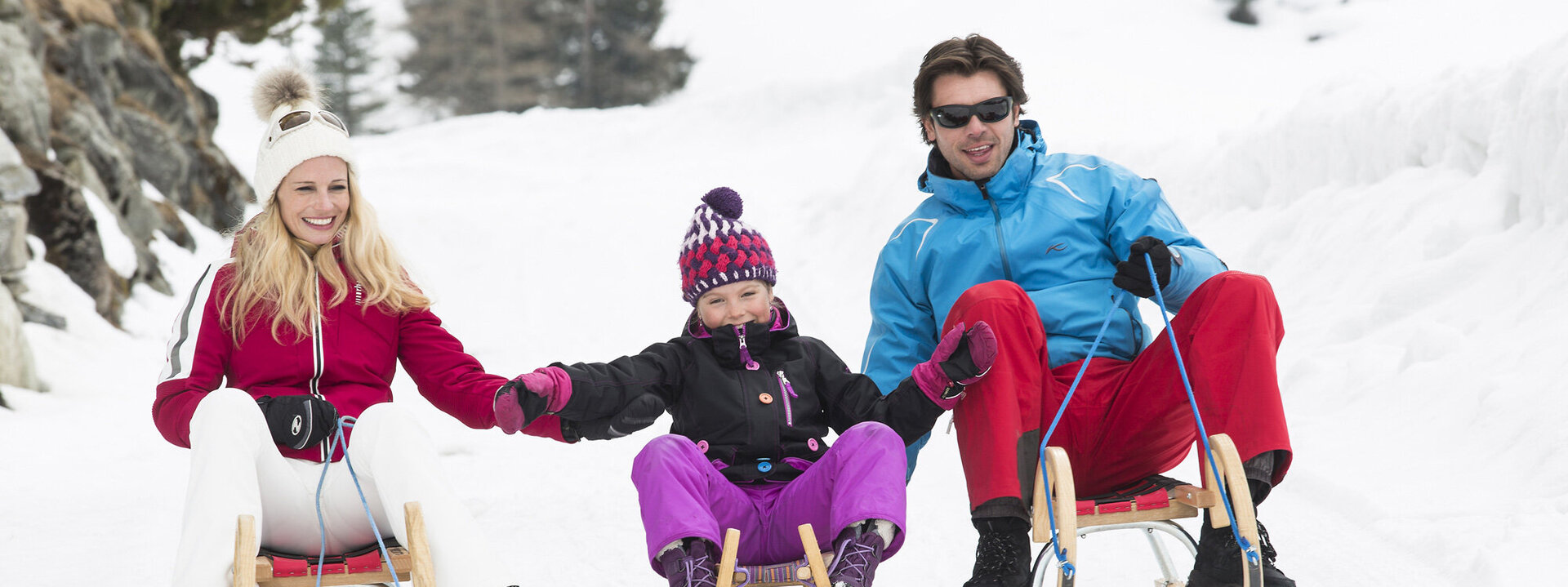 Eine fröhliche Familie sitzt auf Schlitten im Schnee, während sie sich an den Händen halten und gemeinsam lachen.