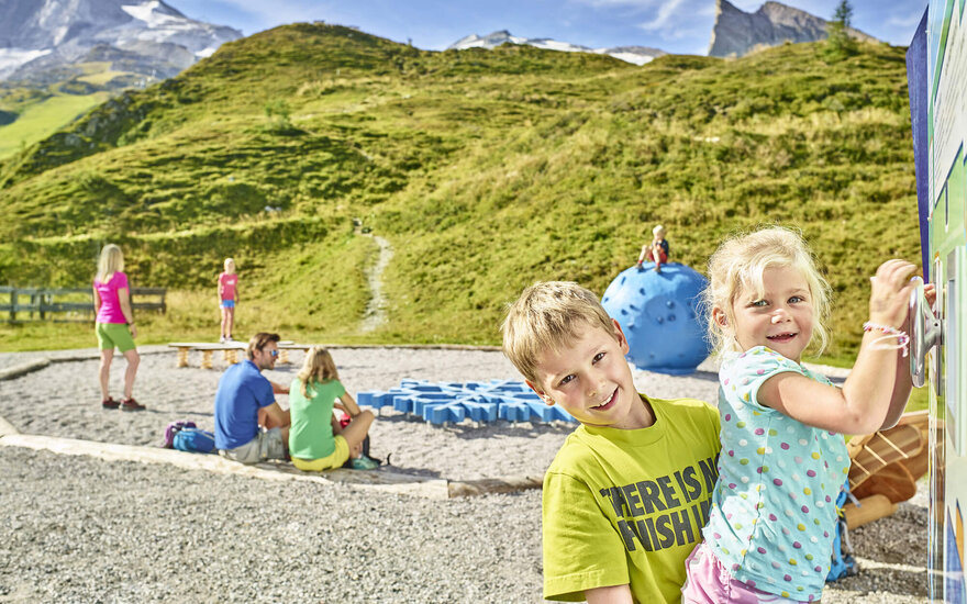 Zwei Kinder spielen fröhlich auf einem Spielplatz in den Bergen, umgeben von grünen Hügeln und blauen Spielgeräten.