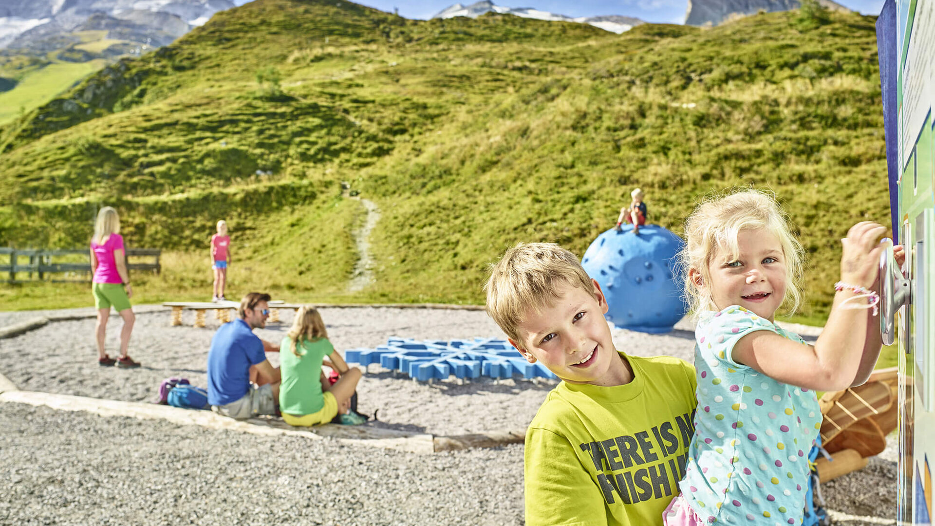 Zwei Kinder spielen fröhlich auf einem Spielplatz in den Bergen, umgeben von grünen Hügeln und blauen Spielgeräten.