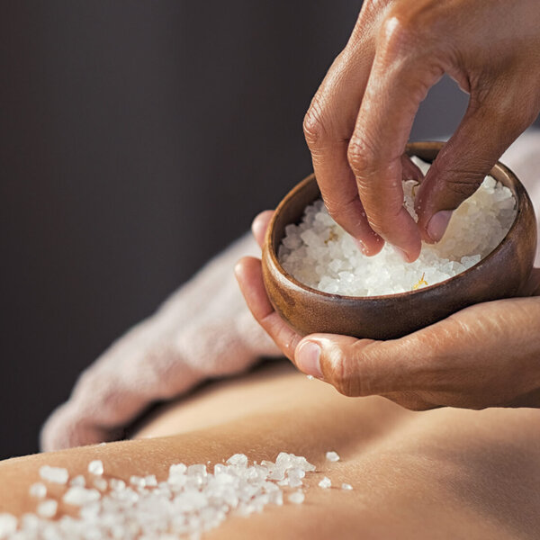 Eine Hand streut grobes Salz aus einer Holzschale auf den Rücken einer Person, die entspannt auf einer Liege liegt.