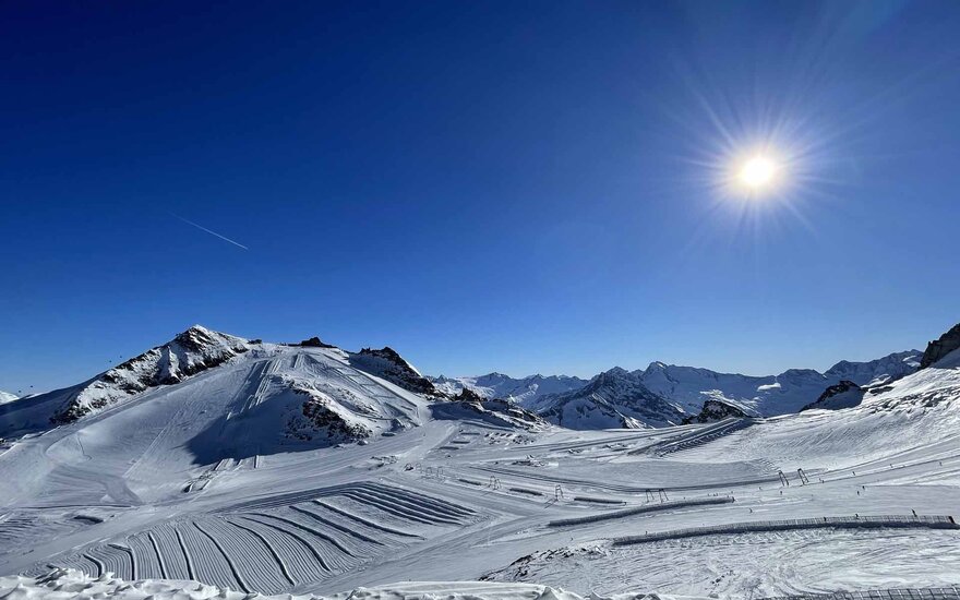 Ein strahlend blauer Himmel über schneebedeckten Bergen, mit der Sonne, die hell am Horizont scheint.