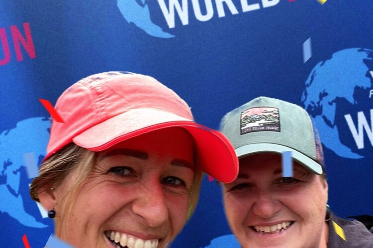 Zwei Frauen lächeln fröhlich in die Kamera, umgeben von bunten Konfetti und einem Hintergrund mit dem Logo des Wings for Life World Run.