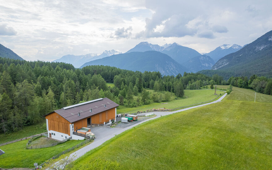Das Bild zeigt eine ländliche Szenerie mit einem modernen Bauernhaus, umgeben von dichten Wäldern und sanften Hügeln.