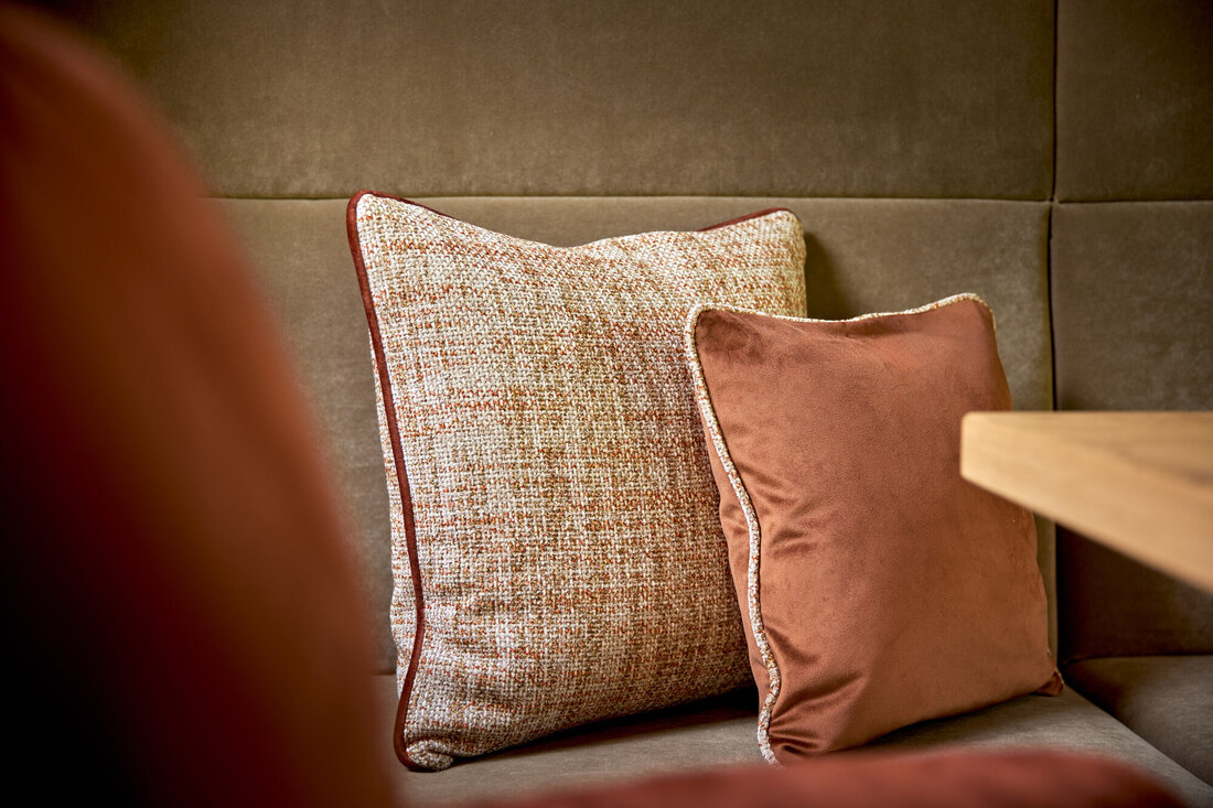 Zwei dekorative Kissen in warmen Erdtönen liegen auf einer beigen Couch, umgeben von einer sanften, neutralen Wand.