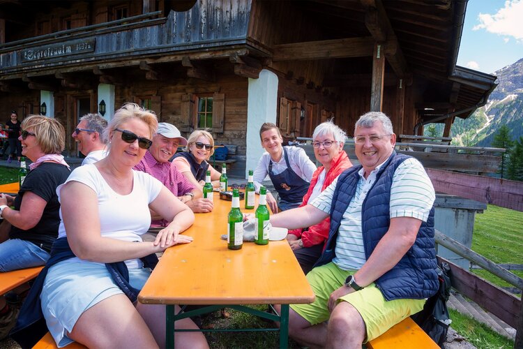 Lächelnde Menschen sitzen an einem Tisch, während sie Getränke genießen und die sonnige Atmosphäre in den Bergen erleben.
