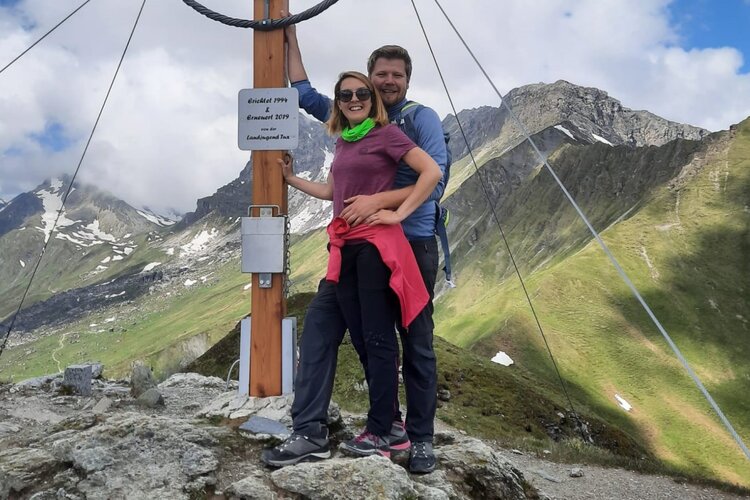 Ein Paar posiert glücklich an einem Berggipfel, umgeben von majestätischen Bergen und grünen Wiesen.