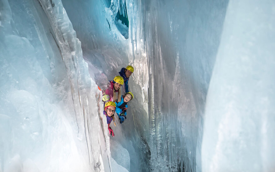 Eine Familie mit Helmen schaut neugierig aus einer Eishöhle, umgeben von glitzerndem Eis und beeindruckenden Eiszapfen.