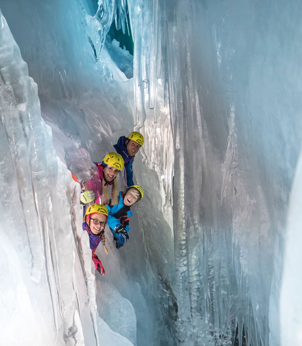 Eine Familie mit Helmen schaut neugierig aus einer Eishöhle, umgeben von glitzerndem Eis und beeindruckenden Eiszapfen.