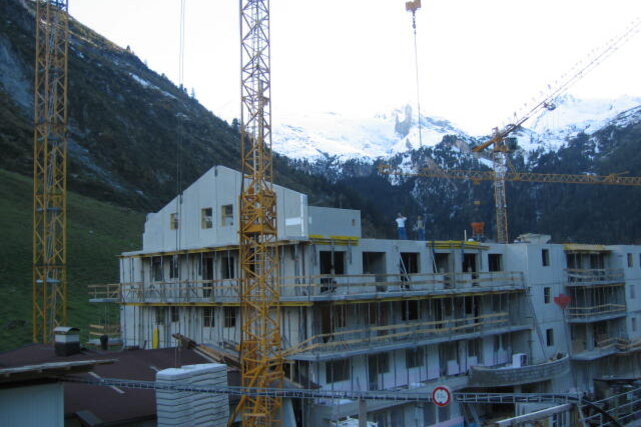 Die Baustelle zeigt Fortschritte beim Bau eines Gebäudes, umgeben von Natur und den schneebedeckten Gipfeln der Alpen.