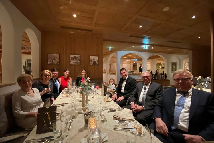 Die Gäste, in formeller Kleidung, genießen ein Abendessen in einem stilvollen, holzverkleideten Raum mit sanfter Beleuchtung.
