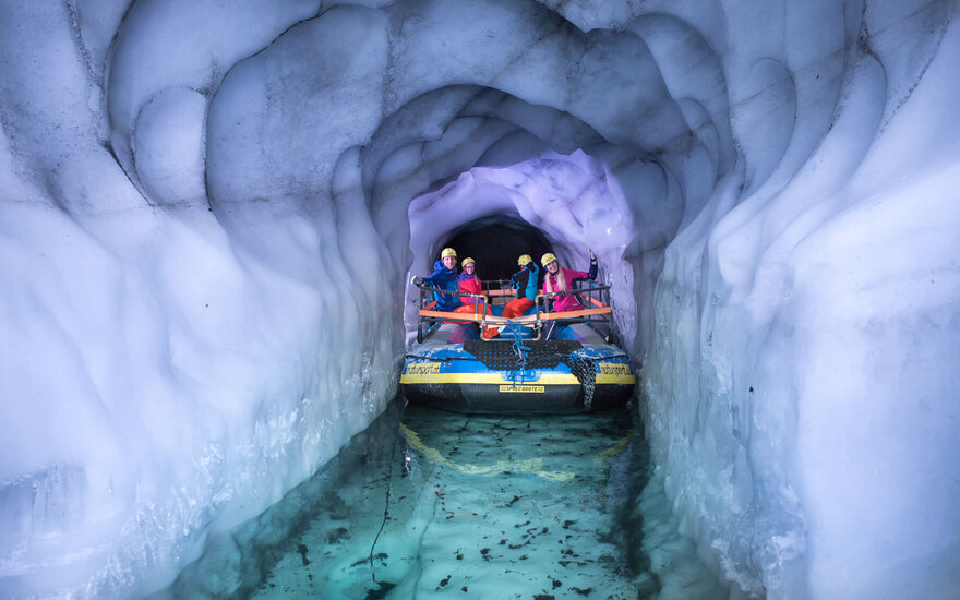 Eine Gruppe von vier Personen in bunten Anzügen sitzt in einem Boot, das durch einen eisigen Tunnel fährt.