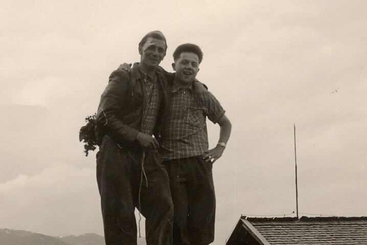 Zwei Männer stehen fröhlich auf einem großen Felsen, umgeben von einer malerischen Berglandschaft und einem Holzhaus.