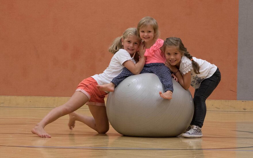 In einer Sporthalle haben drei Mädchen Spaß, während sie auf einem Gymnastikball sitzen und sich gegenseitig umarmen.