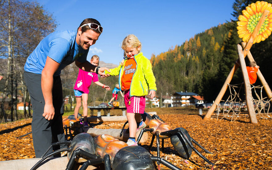 Ein Vater hilft seiner Tochter, über große, bunte Ameisen auf einem Spielplatz zu balancieren, während andere Kinder spielen.