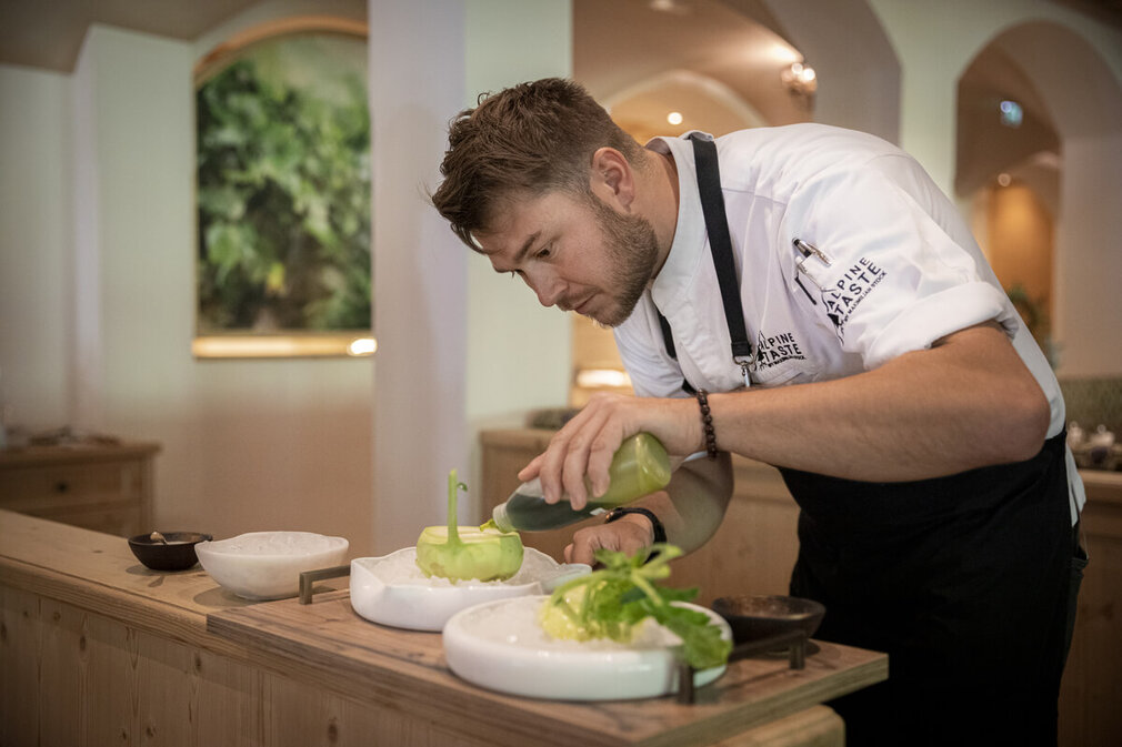 Ein Koch in einer eleganten Küche verfeinert ein Gericht mit einer grünen Sauce, während er konzentriert arbeitet.