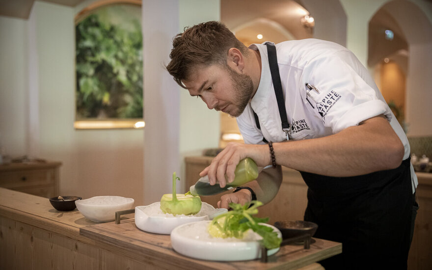 Ein Koch in einer eleganten Küche verfeinert ein Gericht mit einer grünen Sauce, während er konzentriert arbeitet.
