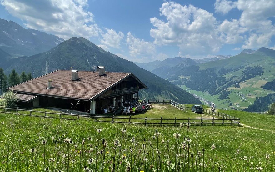 Eine Berghütte steht inmitten einer grünen Wiese, umgeben von majestätischen Bergen und einem strahlend blauen Himmel.