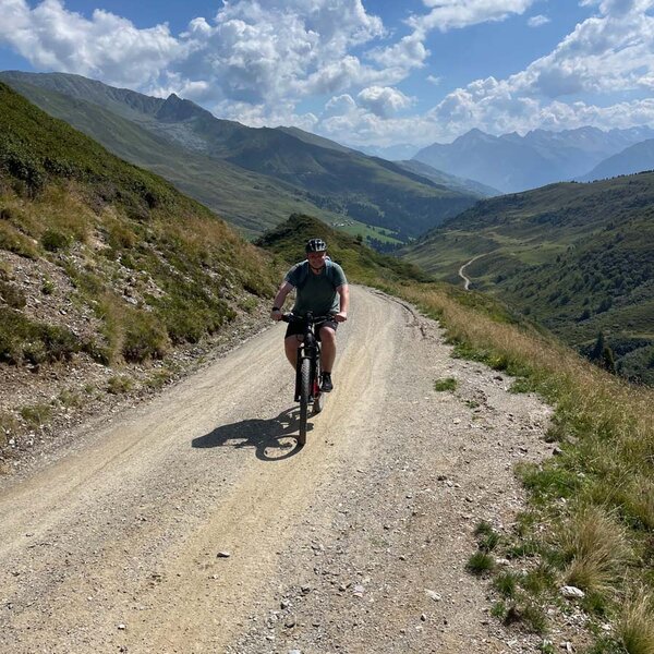 Ein Radfahrer fährt auf einem schmalen Schotterweg, umgeben von grünen Hügeln und majestätischen Bergen im Hintergrund.