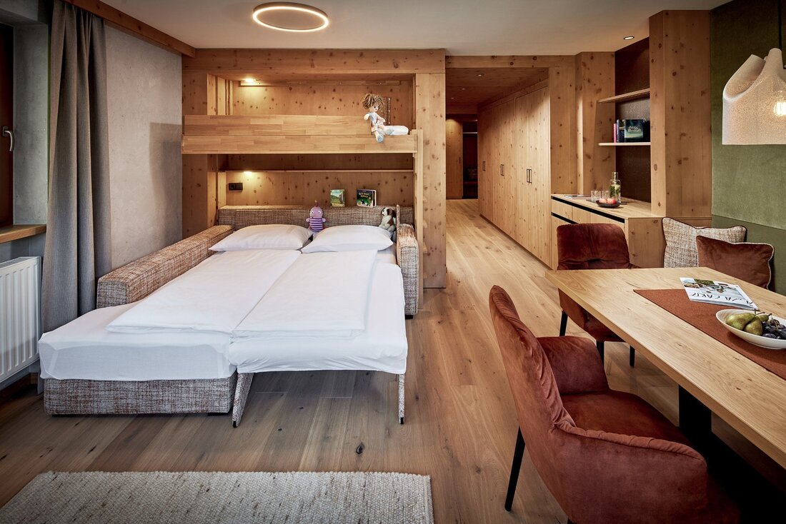 Ein gemütlicher Raum mit einem Schlafsofa, einem Holzbett und einem Esstisch, umgeben von warmen Holztönen und sanftem Licht.