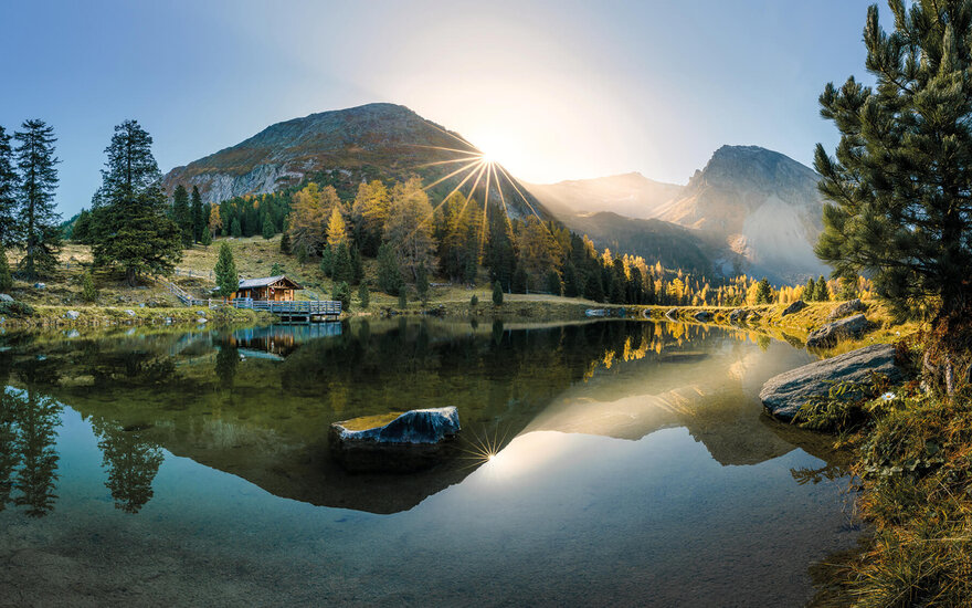 Ein ruhiger Bergsee spiegelt die majestätischen Berge und die strahlende Sonne am Horizont wider, umgeben von buntem Laub.