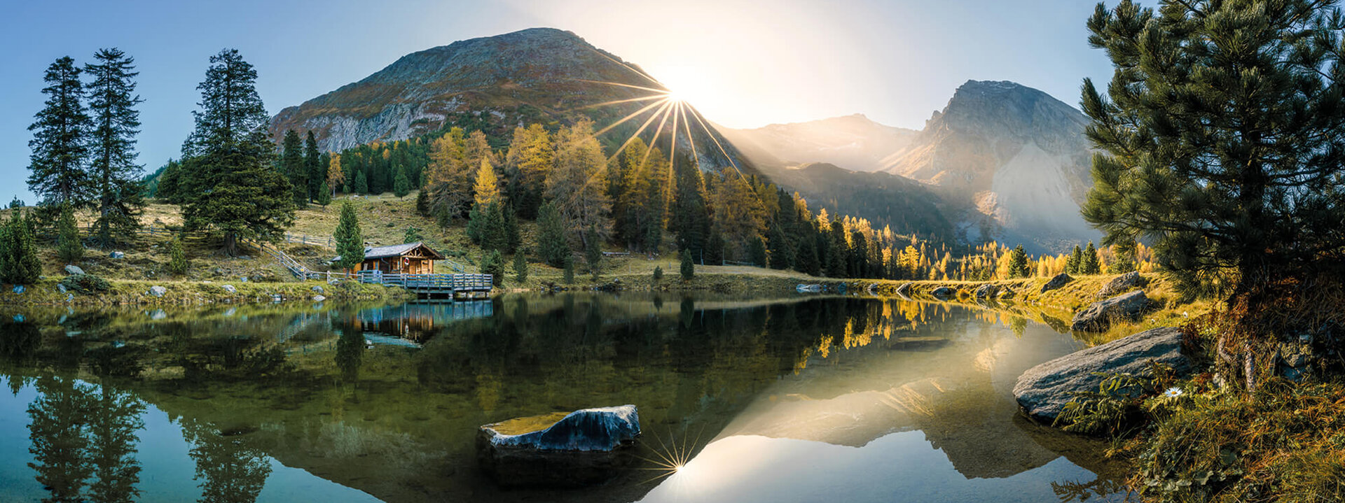 Ein ruhiger Bergsee spiegelt die majestätischen Berge und die strahlende Sonne am Horizont wider, umgeben von buntem Laub.