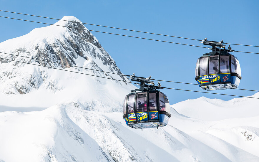 Zwei moderne Gondeln schweben über schneebedeckte Berge, während der klare blaue Himmel im Hintergrund strahlt.