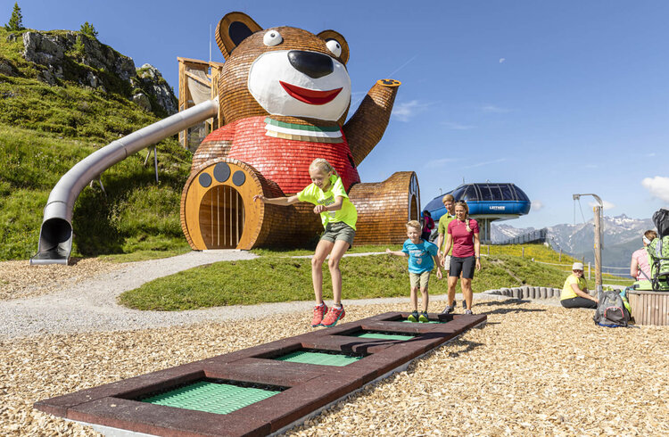 Ein großer, freundlicher Bären-Spielplatz mit einer Rutsche und Kindern, die fröhlich auf einem Trampolin springen.