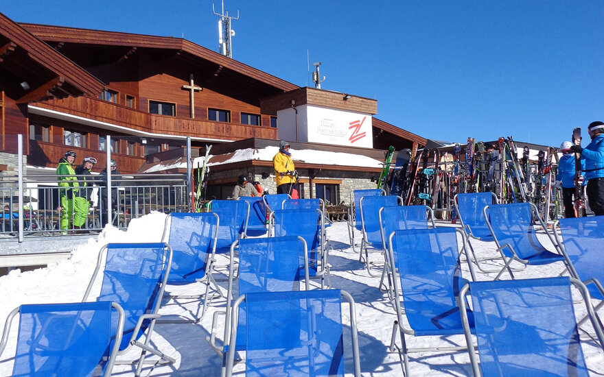 Eine Reihe blauer Liegestühle steht im Schnee vor einer Berghütte unter strahlend blauem Himmel.
