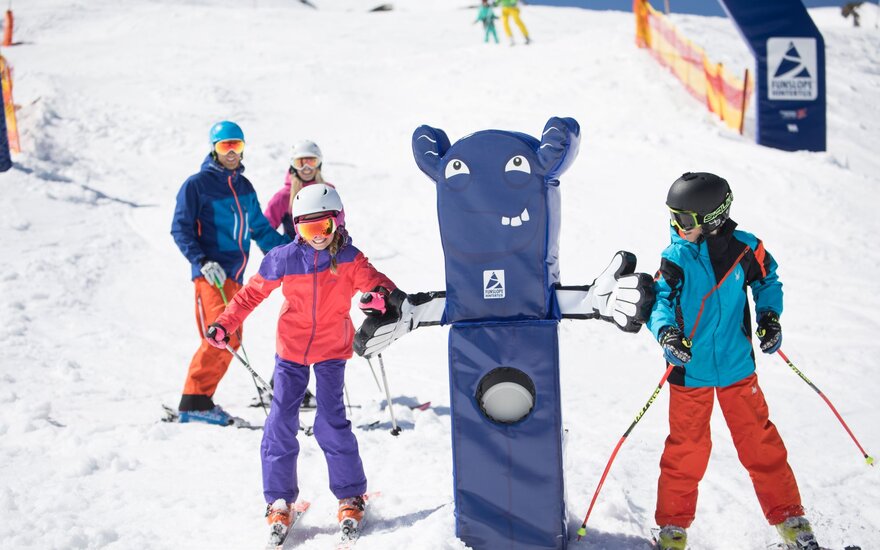 Eine Gruppe von Skifahrern posiert fröhlich im Schnee, während ein lustiger Skimaskottchen mit ihnen interagiert.