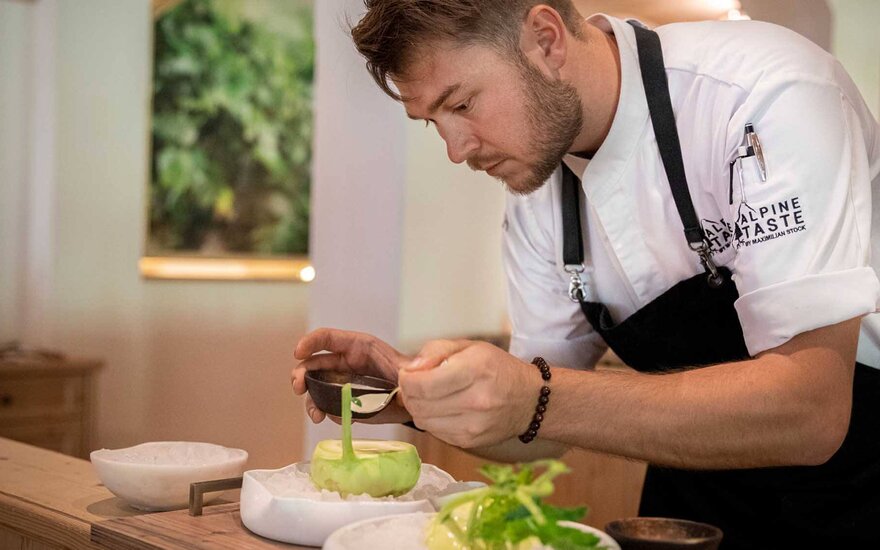 Ein Koch dekoriert sorgfältig ein Gericht mit frischem Kohlrabi und einer grünen Sauce in einem eleganten Restaurant.