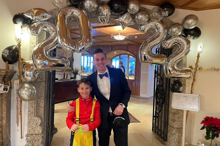 Ein fröhlicher Junge in rotem Shirt und ein Mann in Anzug stehen unter einer Luftballon-Dekoration mit der Zahl 2022.