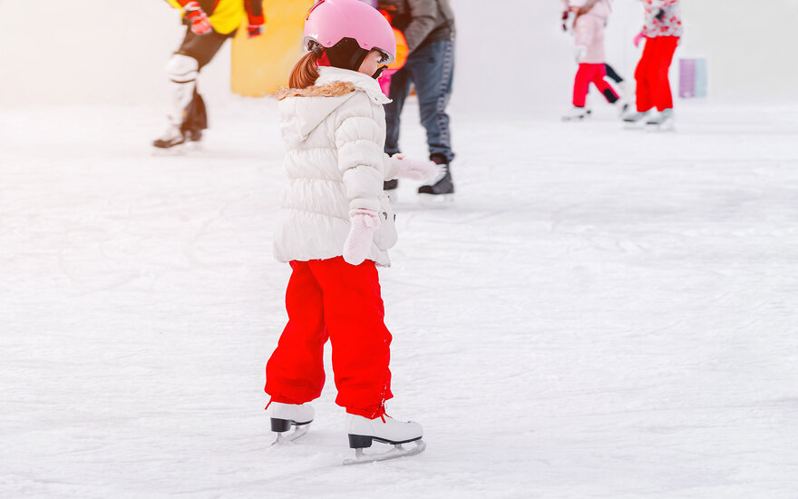 Ein Mädchen mit einem rosa Helm und einer weißen Jacke übt das Eislaufen auf einer glatten Eisfläche.