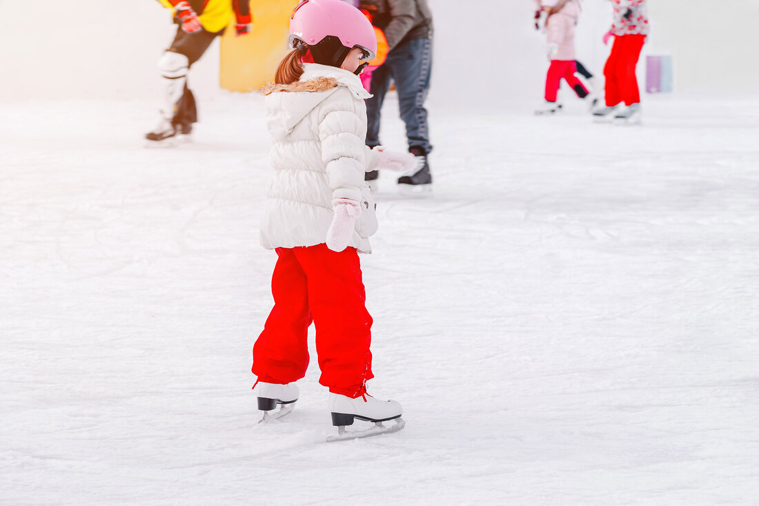 Ein Mädchen mit einem rosa Helm und einer weißen Jacke übt das Eislaufen auf einer glatten Eisfläche.