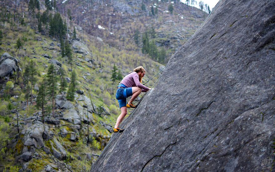 Eine Person klettert an einer steilen Felswand in einer grünen, bewaldeten Berglandschaft.